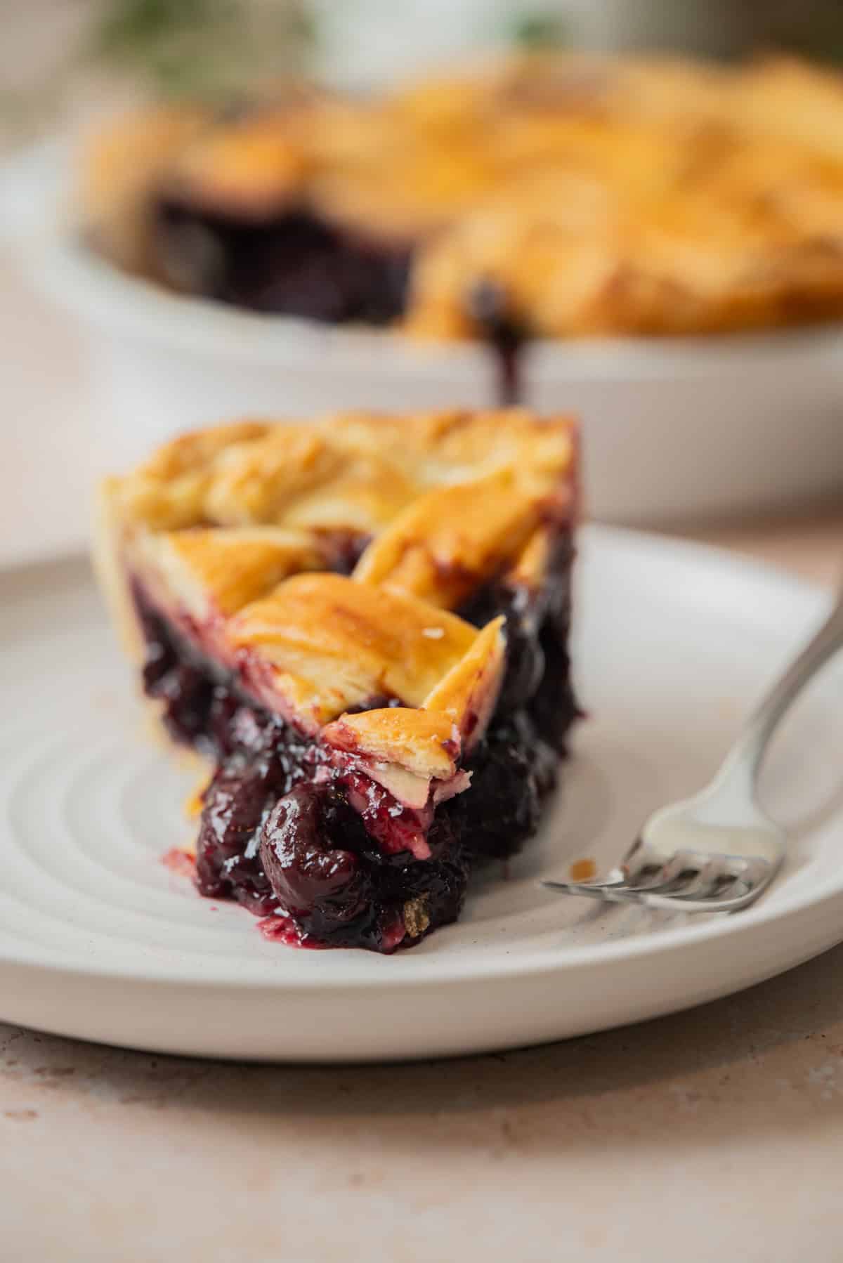 A close-up of a slice of blueberry pie with a golden lattice crust, served on a white plate with a fork. The juicy, vibrant filling looks as delicious as Cherry Pie made with frozen cherries, with the rest of the pie visible in the background.