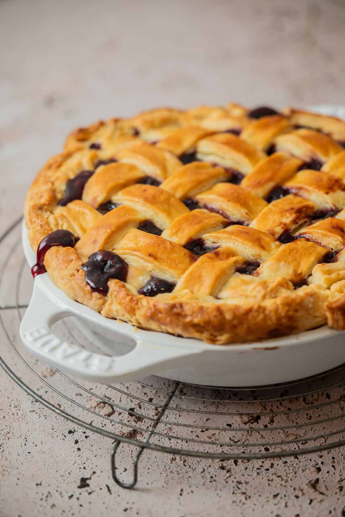 A freshly baked cherry pie with frozen cherries boasts a golden-brown lattice crust, sitting in a white ceramic dish on a cooling rack, with dark fruit filling peeking through the crust strips.