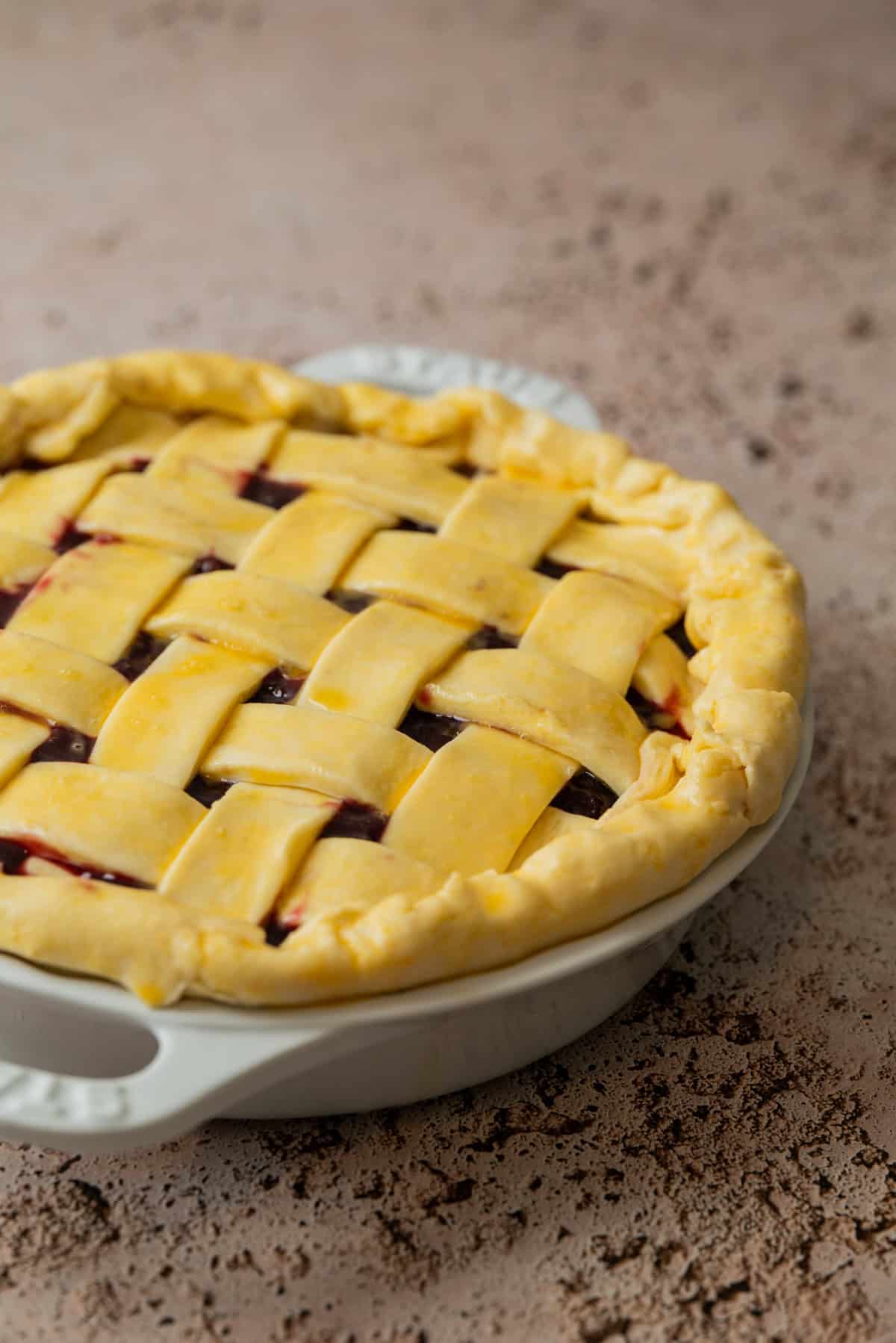 A homemade cherry pie with frozen cherries peeks through a golden, lattice-patterned crust, resting in a white ceramic pie dish on a textured, light brown surface. Some vibrant filling is visible through the lattice.