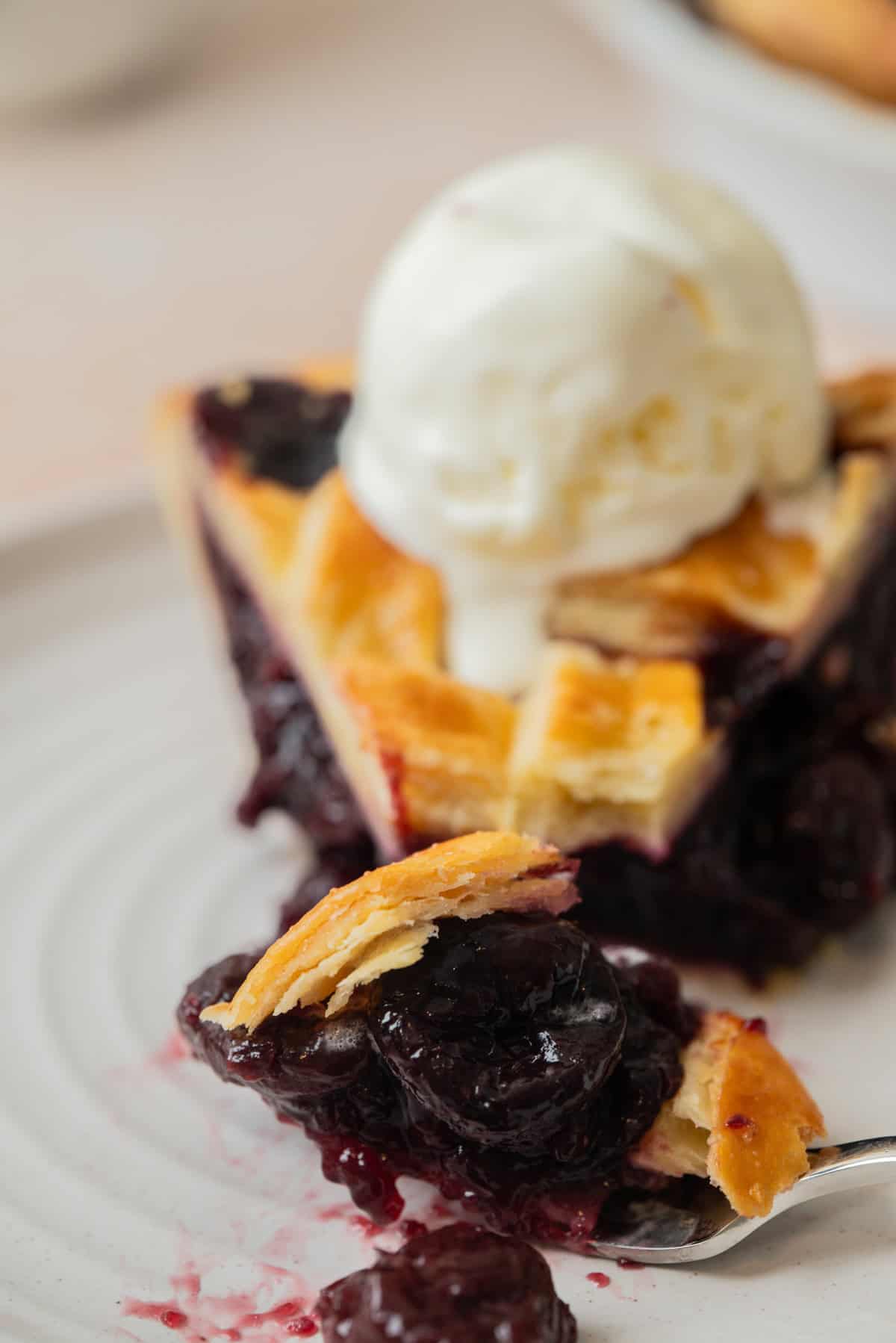 A close-up of a slice of Cherry Pie with Frozen Cherries, topped with a scoop of vanilla ice cream on a white plate. A fork holds a bite of the flaky crust and cherry filling in the foreground.