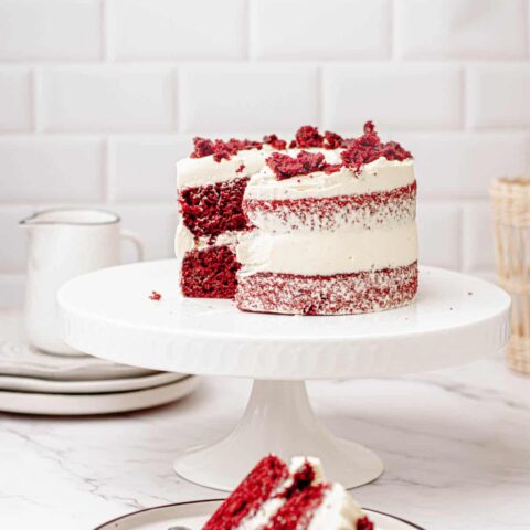 A 6-inch red velvet cake with cream cheese frosting sits elegantly on a white cake stand. A slice is missing, revealing the cake's rich, velvety interior. In the background, there's a small white jug and stacked plates against a tiled wall, completing this picture-perfect scene.