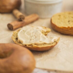 A sliced Pumpkin Bagel with a bite taken out, spread with cinnamon cream cheese, sits on parchment paper alongside two cinnamon sticks and other bagels in the background.