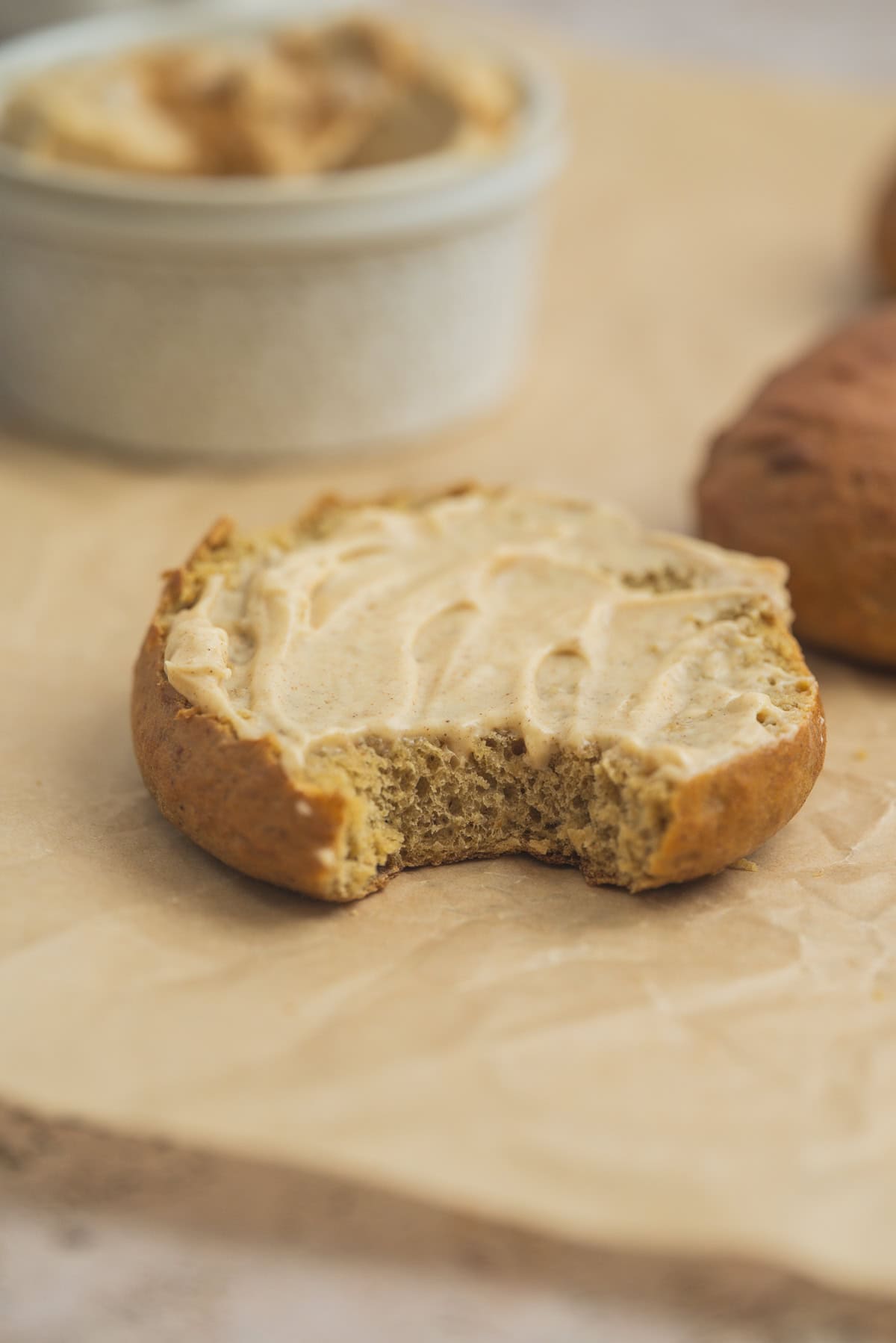 A round Pumpkin Bagel with a bite taken out, topped with a creamy spread, sits on parchment paper. A small dish with more spread and another bagel are blurred in the background.