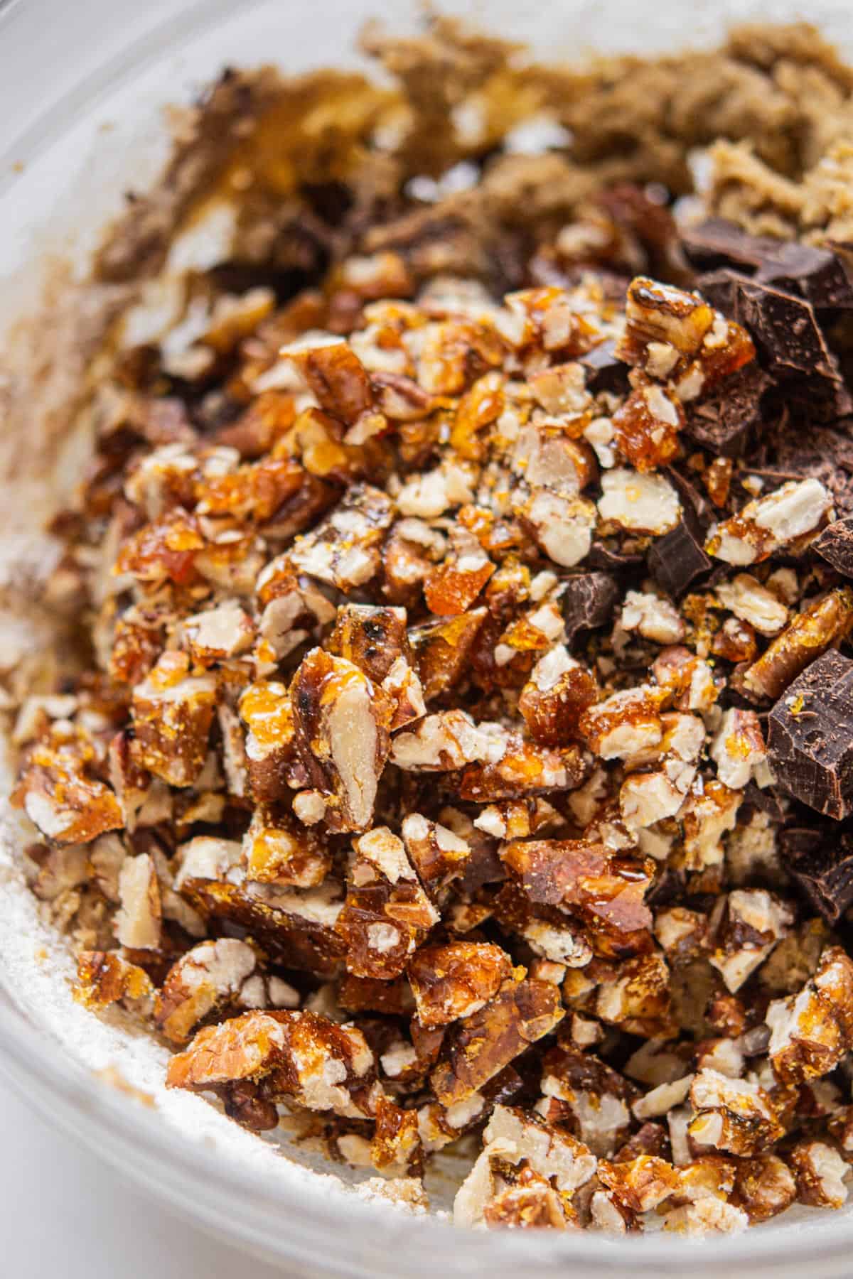 Close-up of a glass bowl filled with chopped candied nuts and dark chocolate chunks—essential ingredients for rich, homemade Chocolate Chip Pecan Cookies—surrounded by classic cookie dough components.