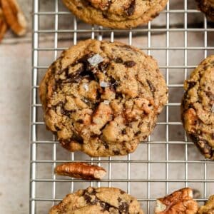 A close-up of Chocolate Chip Pecan Cookies with pecans and a sprinkle of sea salt cooling on a wire rack, with pecan pieces visible nearby.