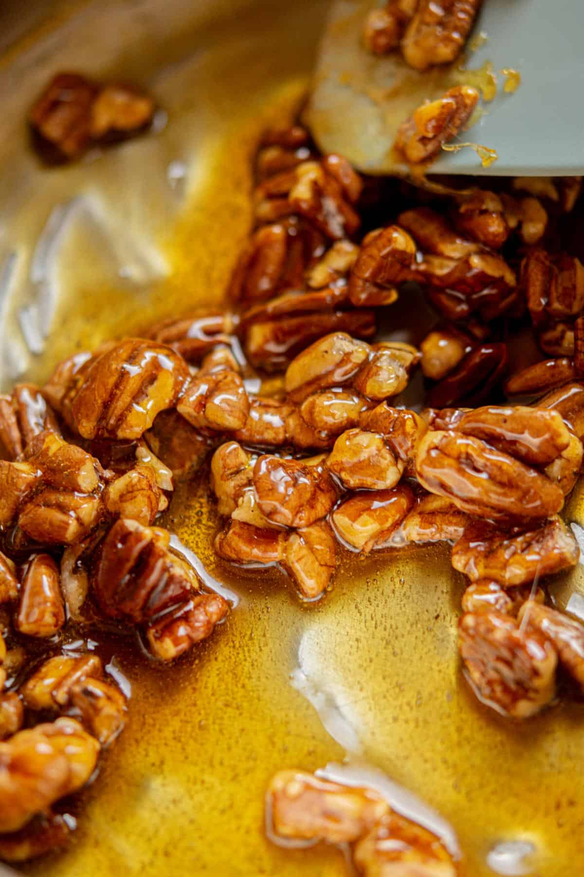 Close-up of toasted pecans coated in glossy caramelized sugar, being stirred with a spatula in a metal pan—essential for making decadent Chocolate Chip Pecan Cookies.