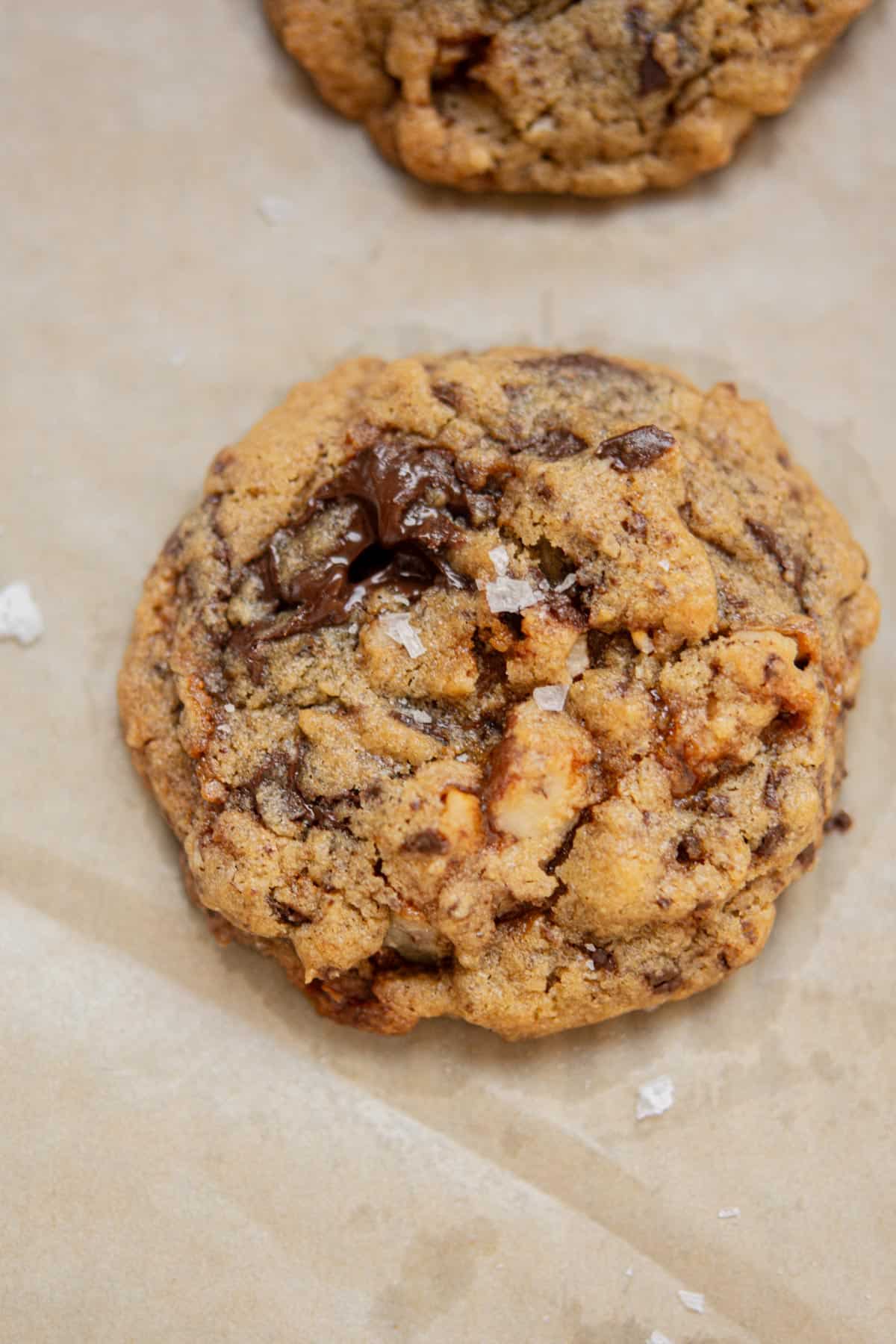 A close-up of a Chocolate Chip Pecan Cookie with visible chocolate chunks, toasted pecans, and a sprinkle of flaky sea salt on top, resting on parchment paper.