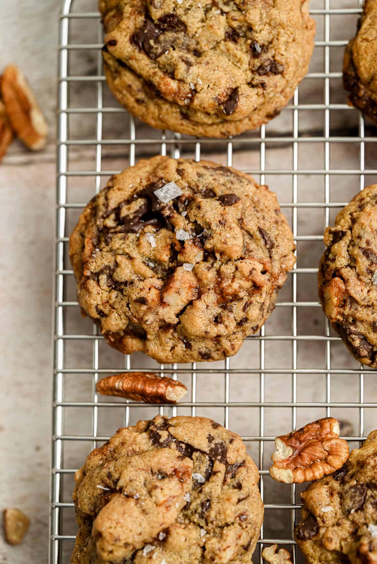 Freshly baked pecan Chocolate Chip Cookies with visible pecans are cooling on a metal wire rack, while a few pecan halves are scattered on the light-colored surface nearby.