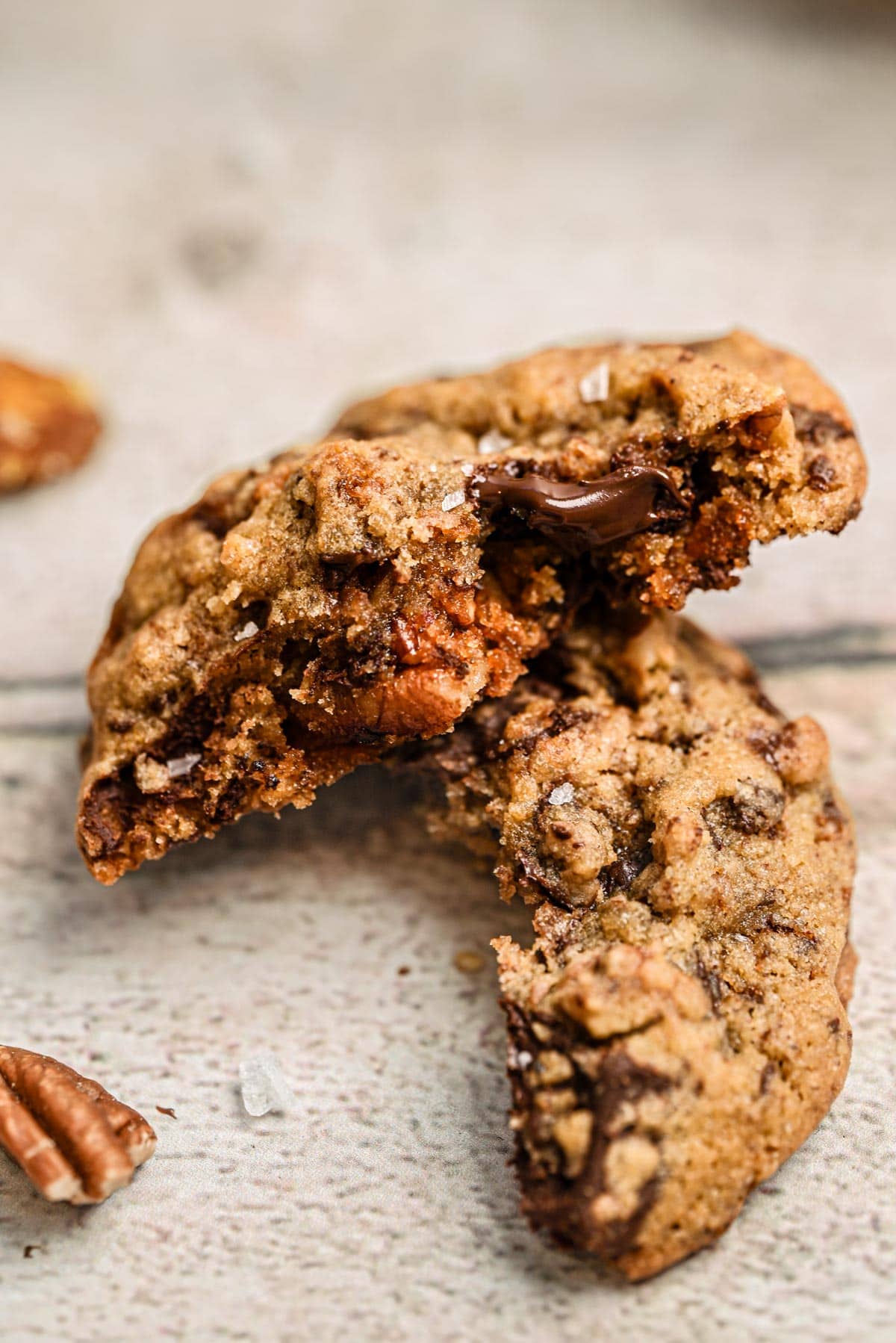 Close-up of Chocolate Chip Pecan Cookies, broken in half to reveal a gooey chocolate center and crunchy pecan pieces, resting on a light-colored textured surface.