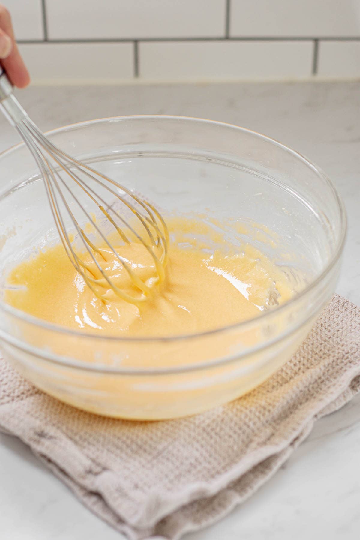 whisked egg yolks in a glass bowl with hand holding a ballon whisk. The bowl is propped on a kitchen towel.