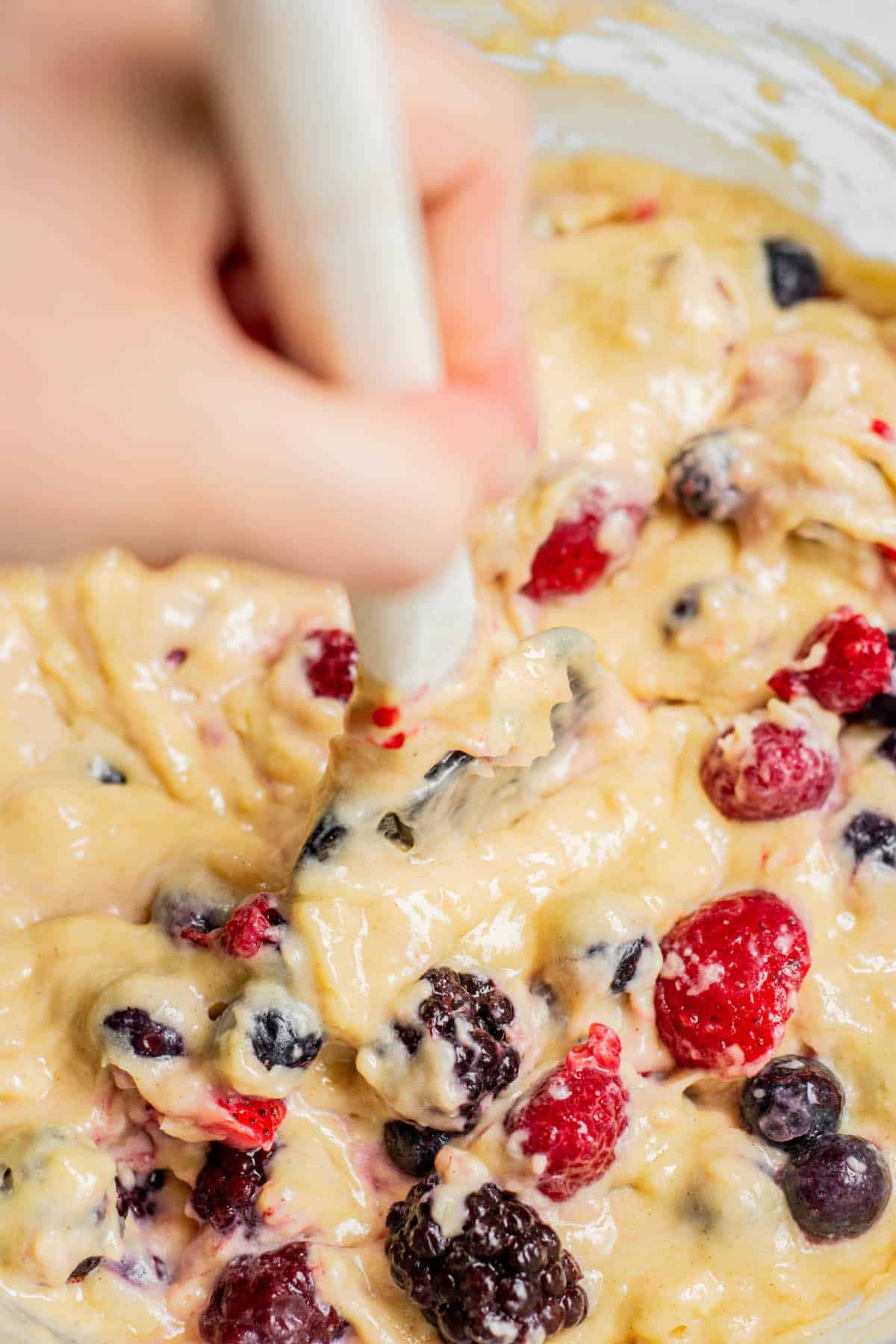 hand holding spatula and folding berries into frozen muffin batter.