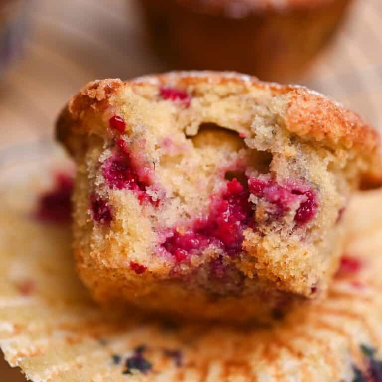 A close-up of a partially eaten Frozen Berry Muffin, showing its moist, crumbly texture and juicy raspberry pieces inside, with the muffin wrapper peeled back.