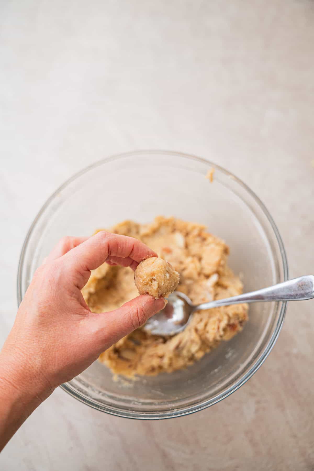 A hand holds a small ball of cookie dough—perfect for making Kletskoppen—over a glass bowl filled with more dough and a spoon, on a light countertop.