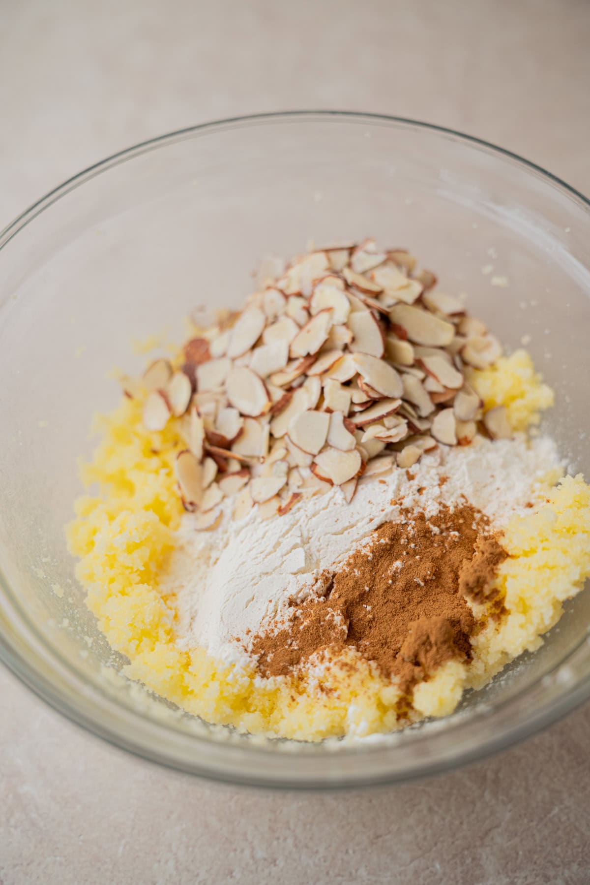 A clear glass bowl containing creamed butter and sugar, topped with sliced almonds, flour, and ground cinnamon—classic ingredients ready to be mixed together for baking delicious Kletskoppen cookies.