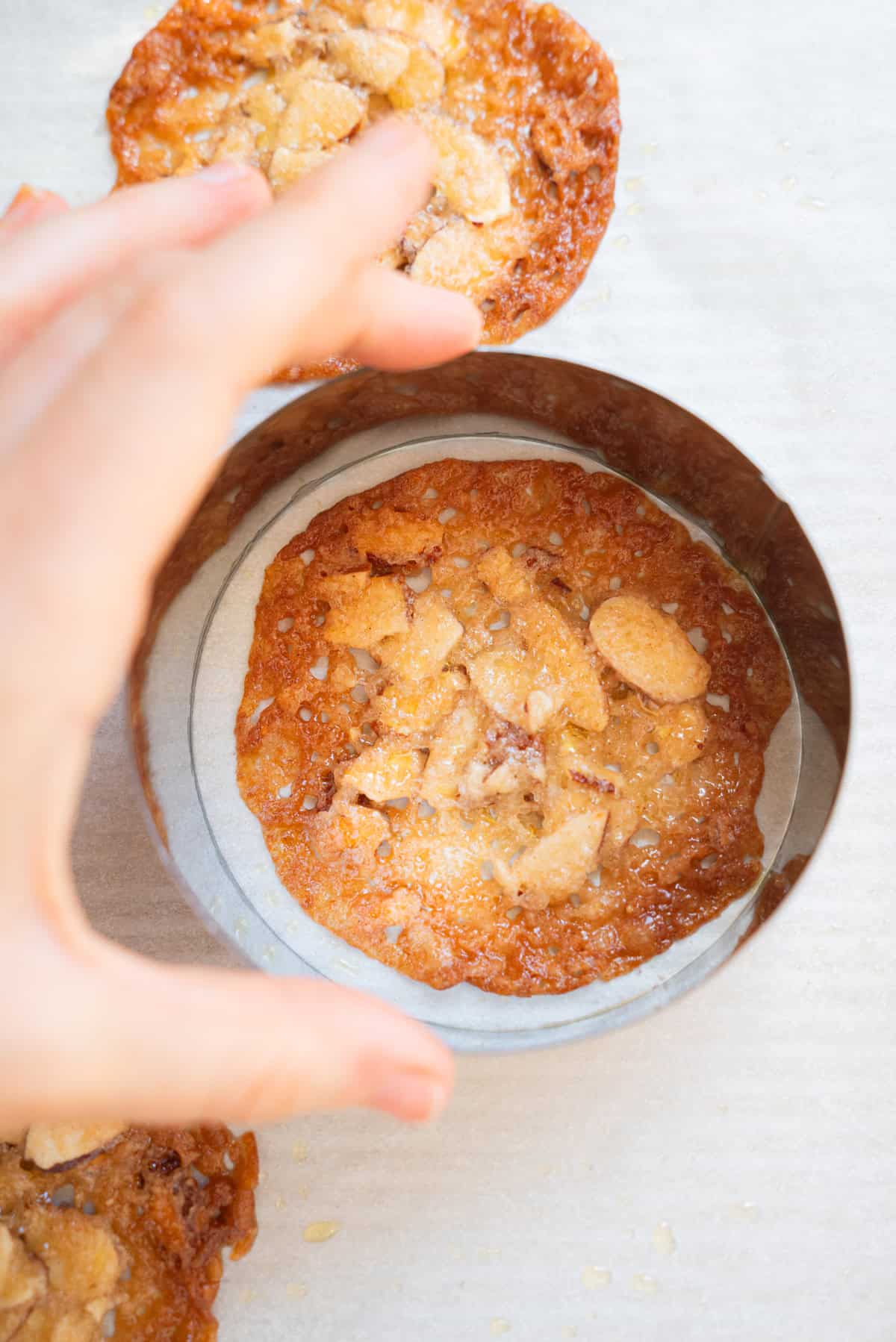 A hand uses a round metal cutter to shape a golden, crispy Kletskoppen cookie with almond pieces on parchment paper.