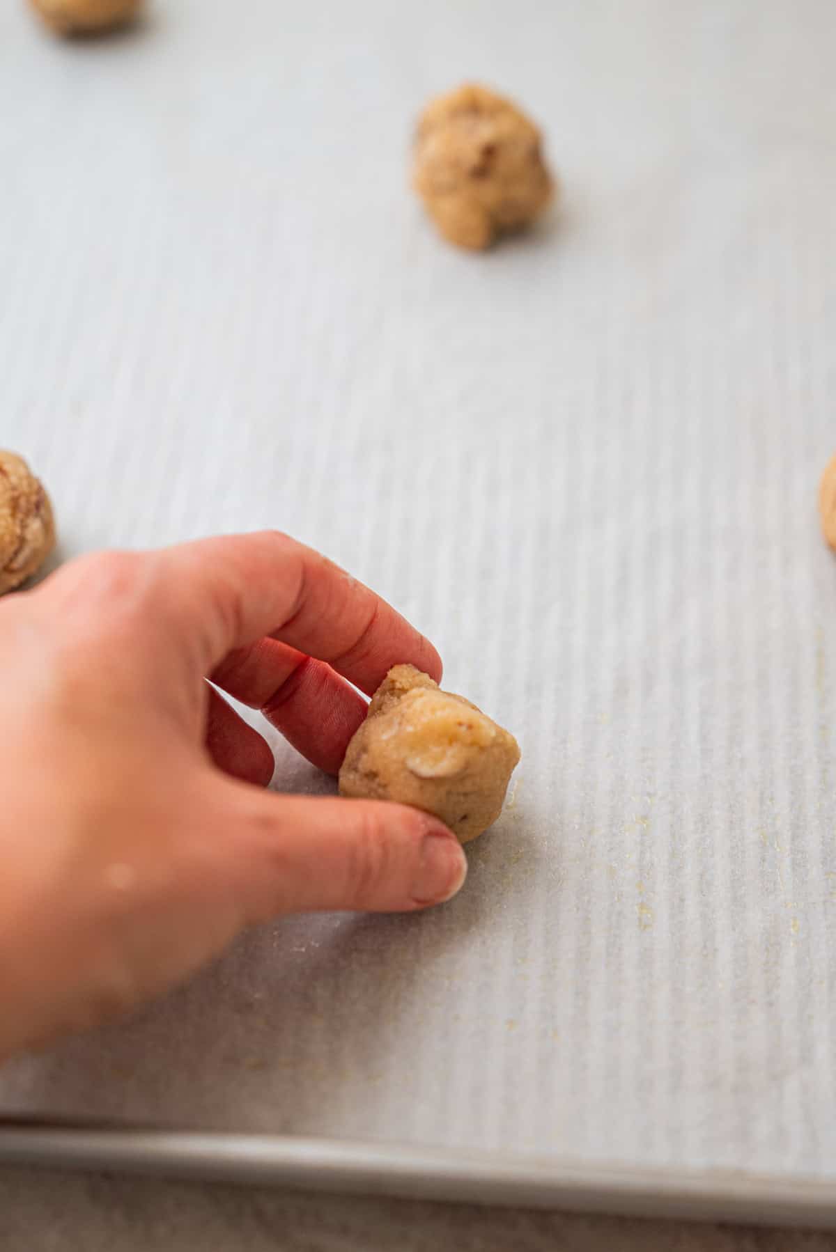 A hand places a ball of cookie dough onto a parchment-lined baking sheet, with other dough balls—including classic Kletskoppen—visible in the background.