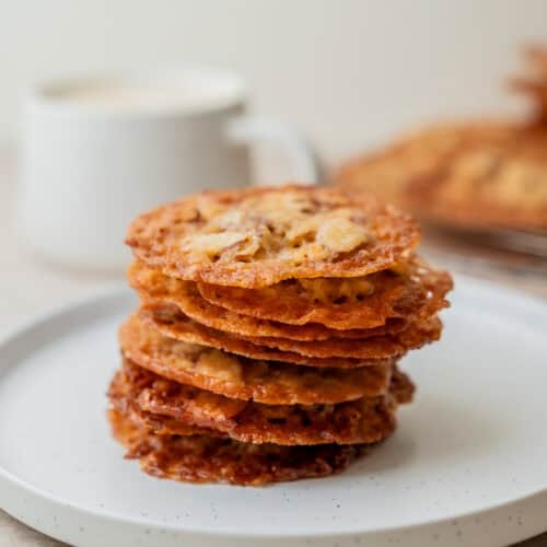 A stack of thin, crispy Kletskoppen cookies sits on a white plate, with a blurred white mug and more cookies on a cooling rack in the background.