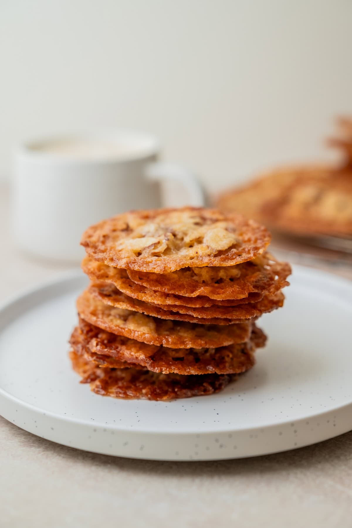 A stack of thin, crispy Kletskoppen cookies sits on a white plate, with a blurred white mug and more cookies on a cooling rack in the background.