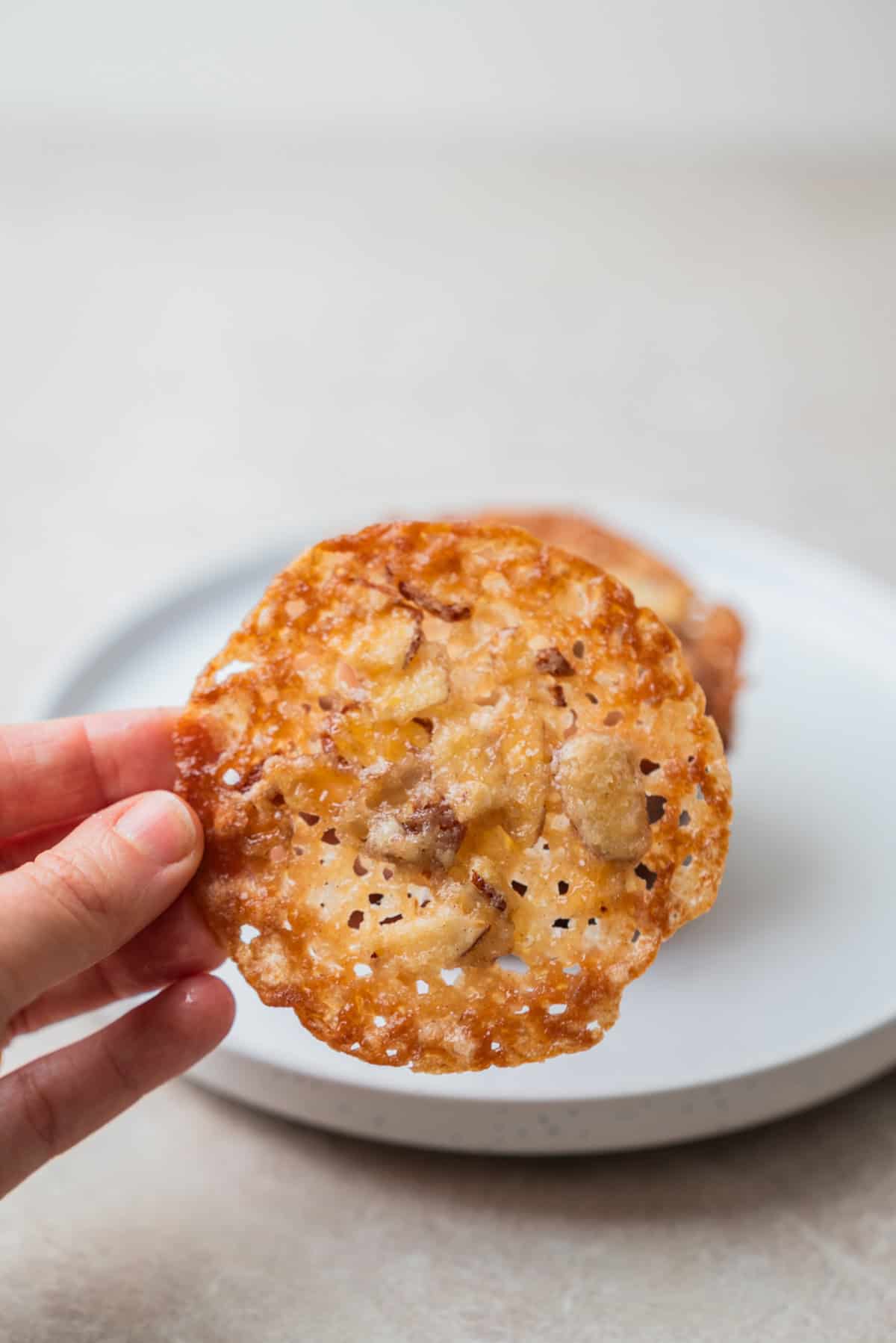 A hand holds up a thin, round, lacy Kletskoppen cookie with visible bits of nuts or toffee, with a blurred white plate and cookie in the background.