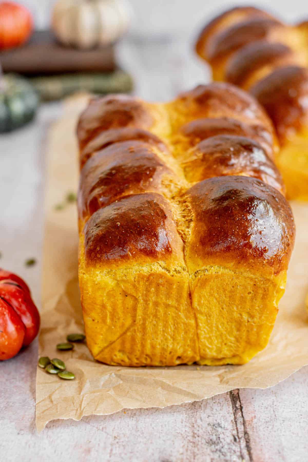 Glistening pumpkin brioche loaf with a golden crust, placed on parchment paper surrounded by scattered pumpkin seeds, with blurred autumnal decorations in the background.