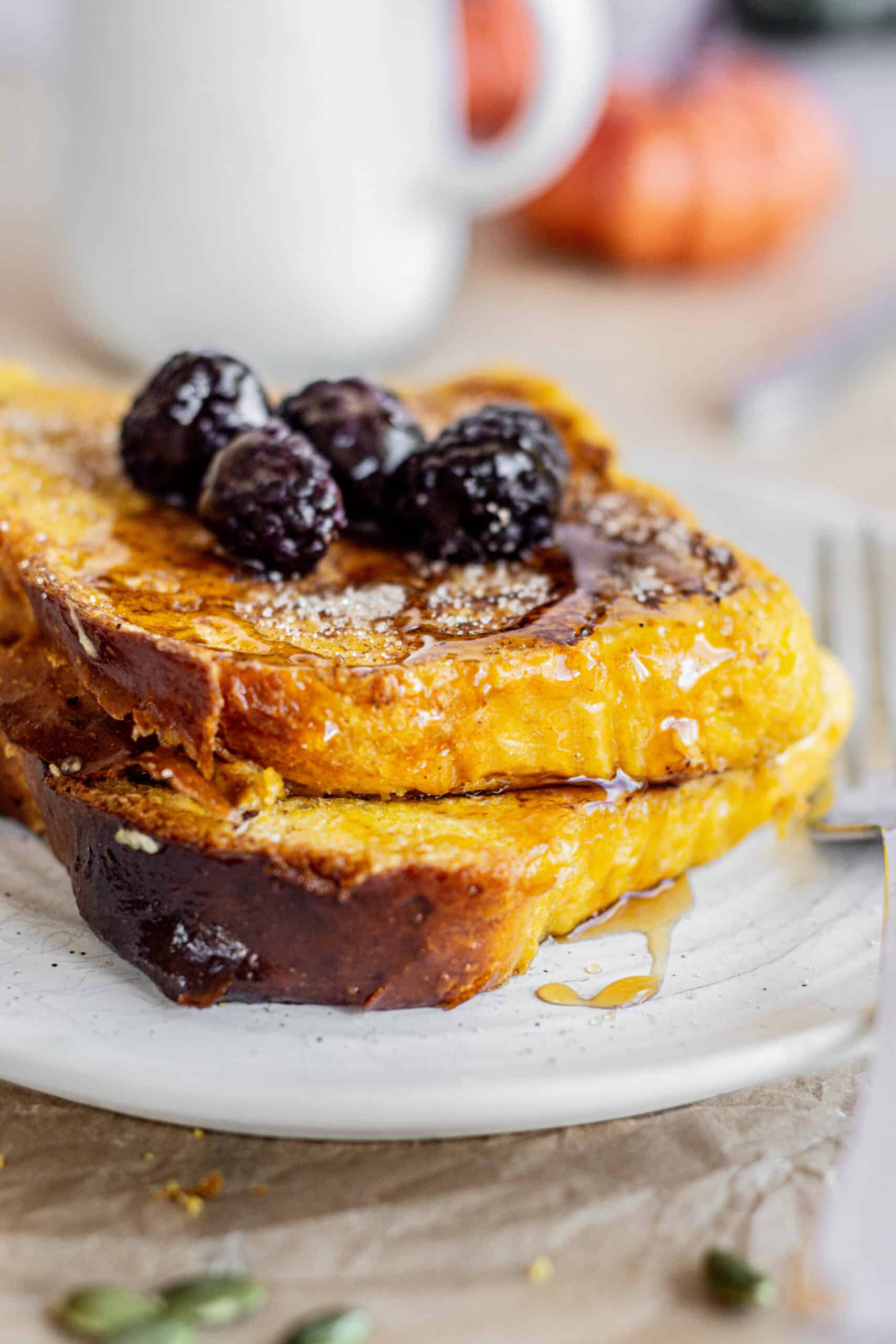 Golden pumpkin French toast topped with glistening blackberries and drizzled syrup, served on a white plate with a blurred white mug in the background.