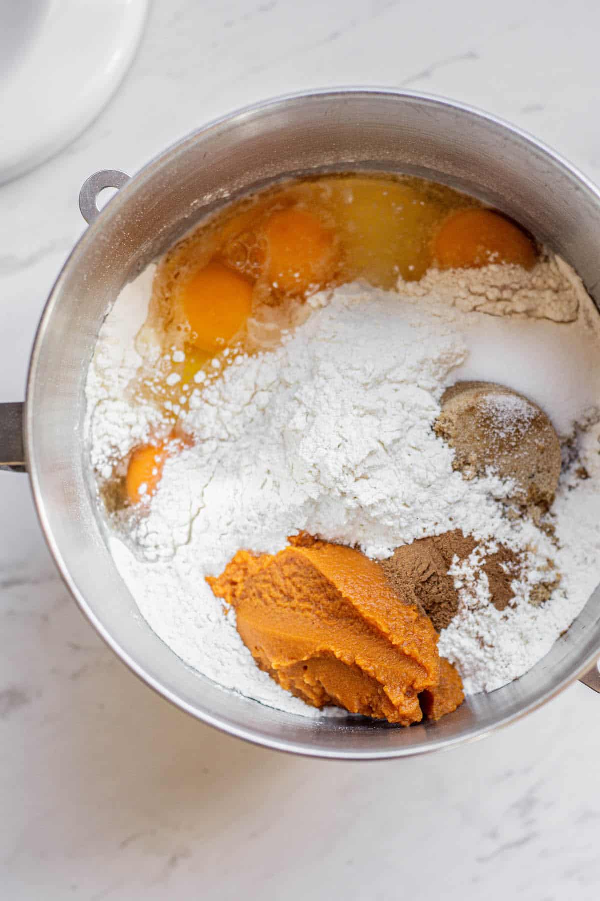 A stainless steel mixing bowl containing ingredients for pumpkin dough: raw eggs, flour, sugar, spices, and a scoop of vibrant pumpkin puree on a white marble surface.