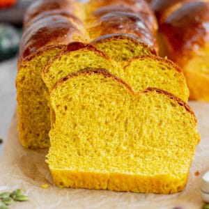 Golden pumpkin brioche loaf with a shiny crust arranged in a staggered pattern. Set on parchment paper with pumpkins, seeds, and a rustic backdrop