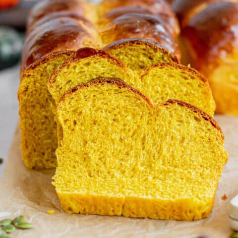 Golden pumpkin brioche loaf with a shiny crust arranged in a staggered pattern. Set on parchment paper with pumpkins, seeds, and a rustic backdrop