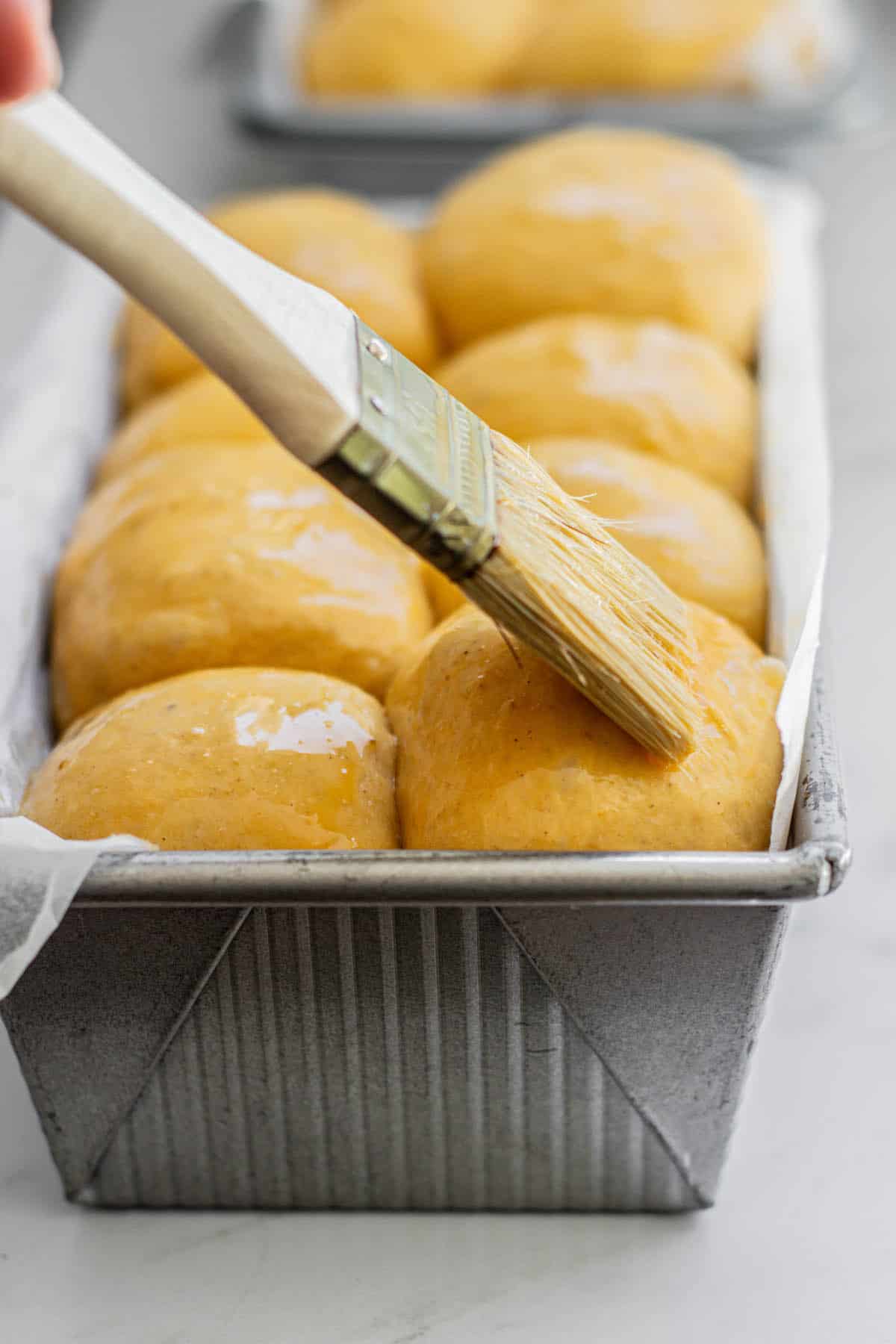 risen golden rolls in a metal baking tin being brushed with egg wash using a pastry brush.