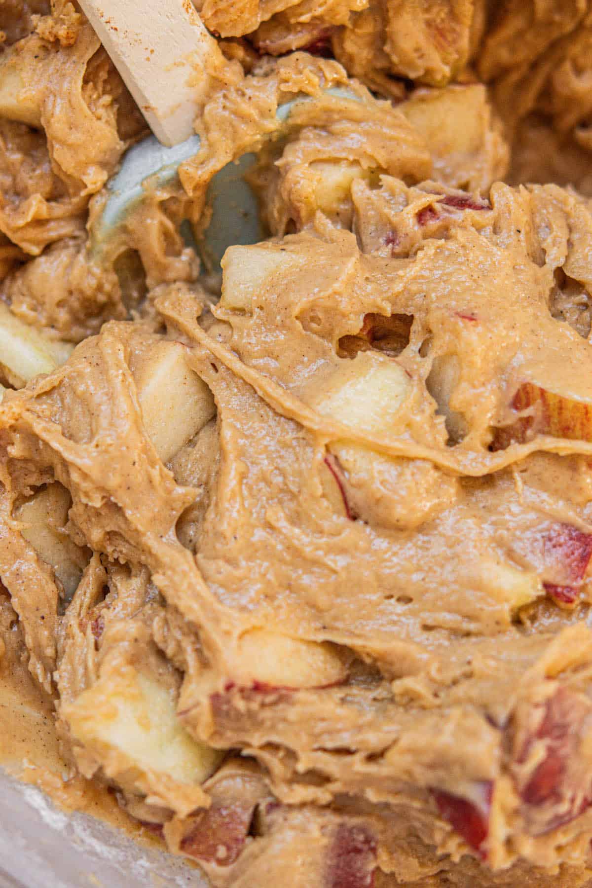 Close-up of thick, brown batter with visible chunks of red-skinned apple being stirred with a light blue spatula.