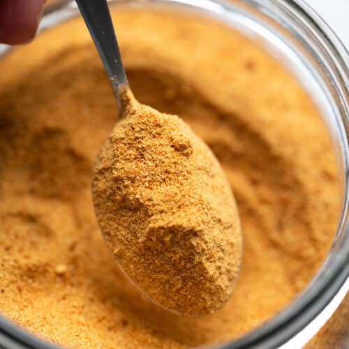 A close-up of a spoonful of orange-brown seasoning powder, reminiscent of toasted milk powder, being scooped from a glass jar.