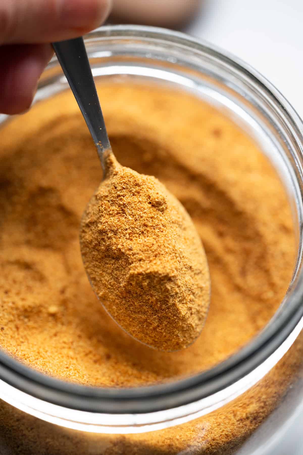 A close-up of a spoonful of orange-brown seasoning powder, reminiscent of toasted milk powder, being scooped from a glass jar.