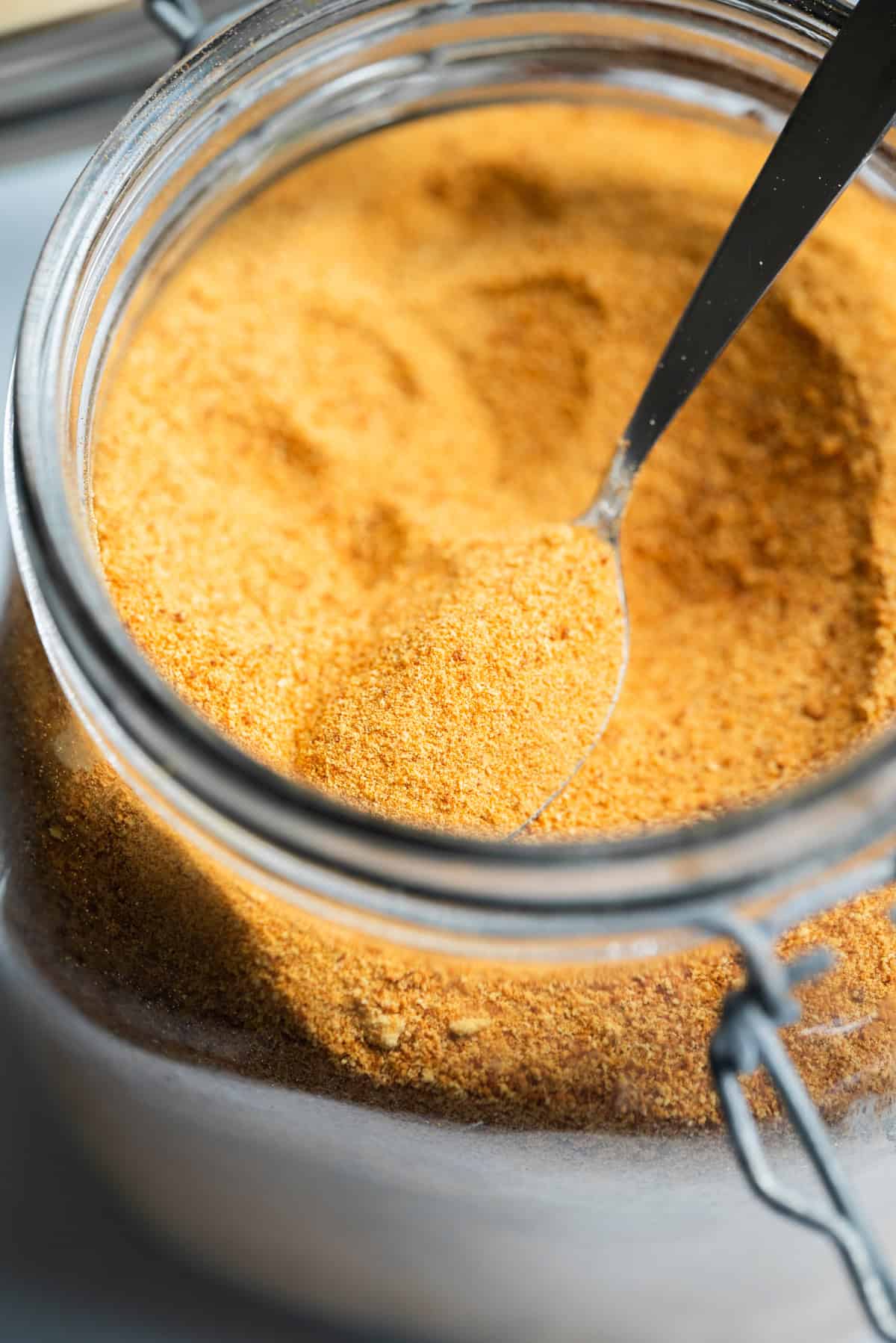 A close-up of a glass jar filled with fine, orange-brown toasted milk powder. A metal spoon is scooping some of the toasted milk powder from the jar. The jar has a hinged lid.