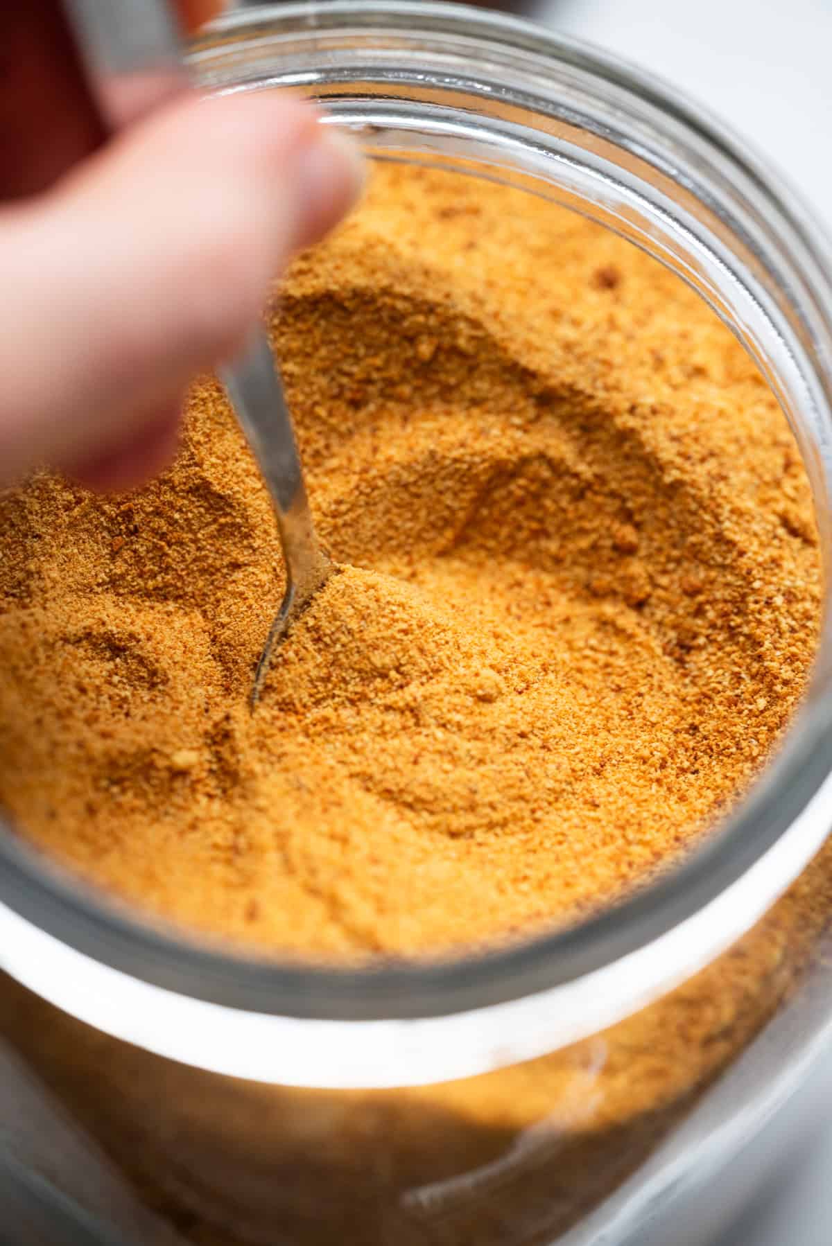 A close-up of a hand holding a spoon, scooping toasted milk powder: its warm orange-brown hue, from a glass jar.