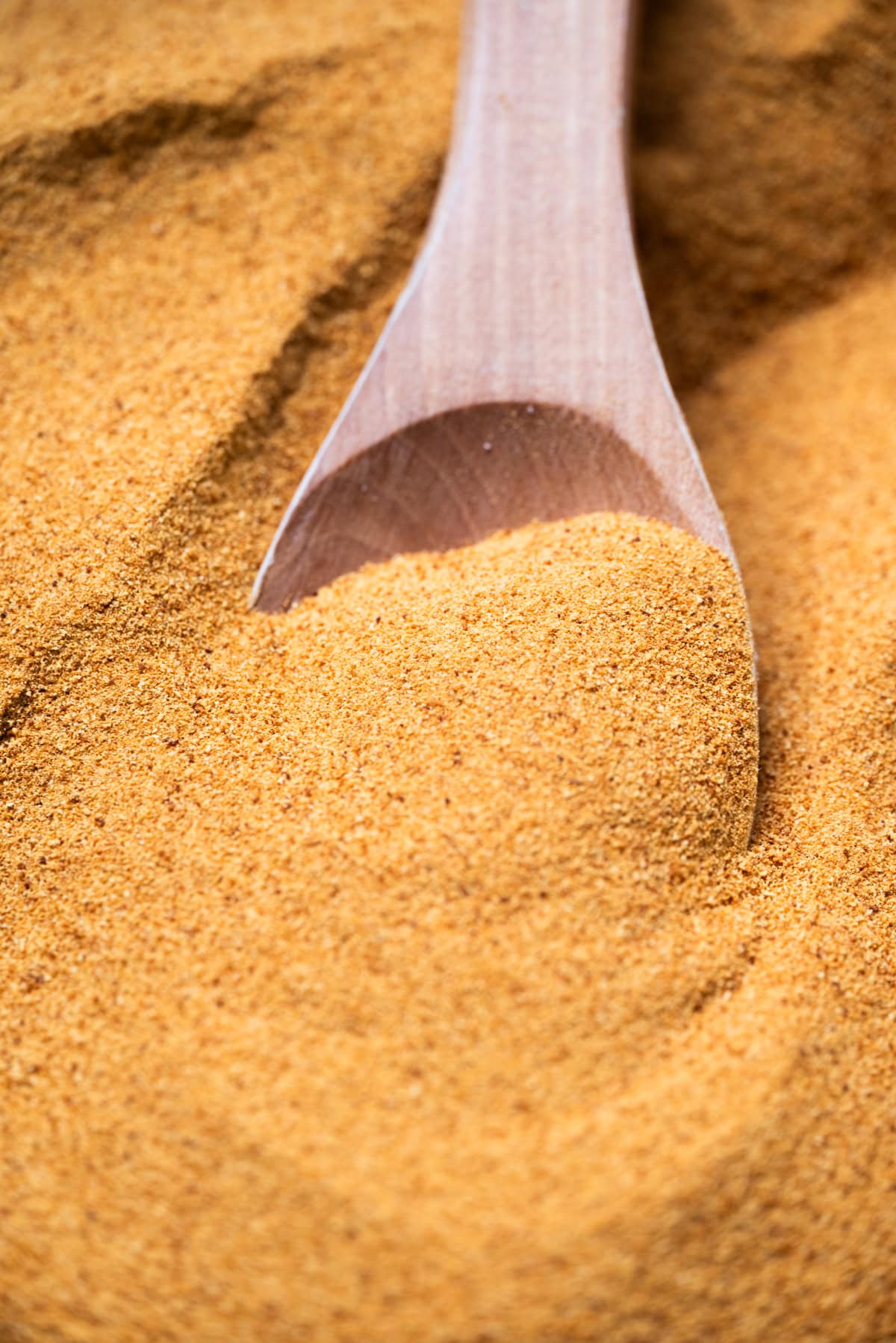A close-up of a wooden spoon scooping fine, golden-brown toasted milk powder from a larger pile of the same powder.