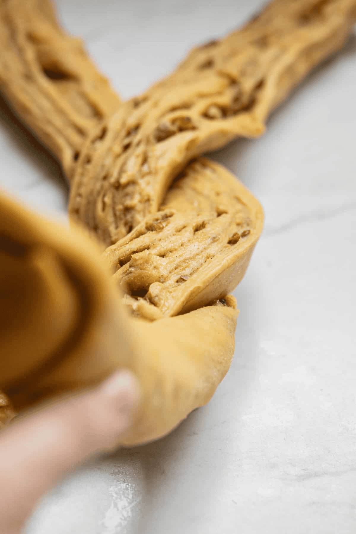 Close-up of a hand braiding three strips of cinnamon-speckled pumpkin babka dough on a light-colored surface, preparing it for baking.