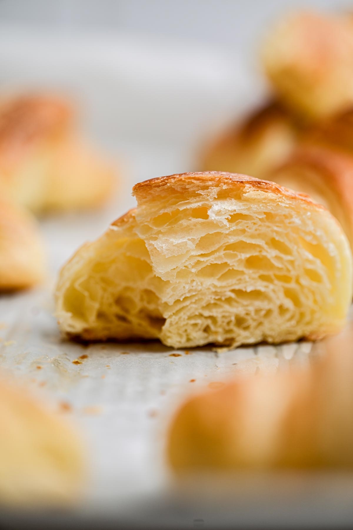 A close-up of a flaky, golden homemade croissant half, showing its airy, layered interior, resting on a white parchment-lined surface with blurred pastry pieces in the background.