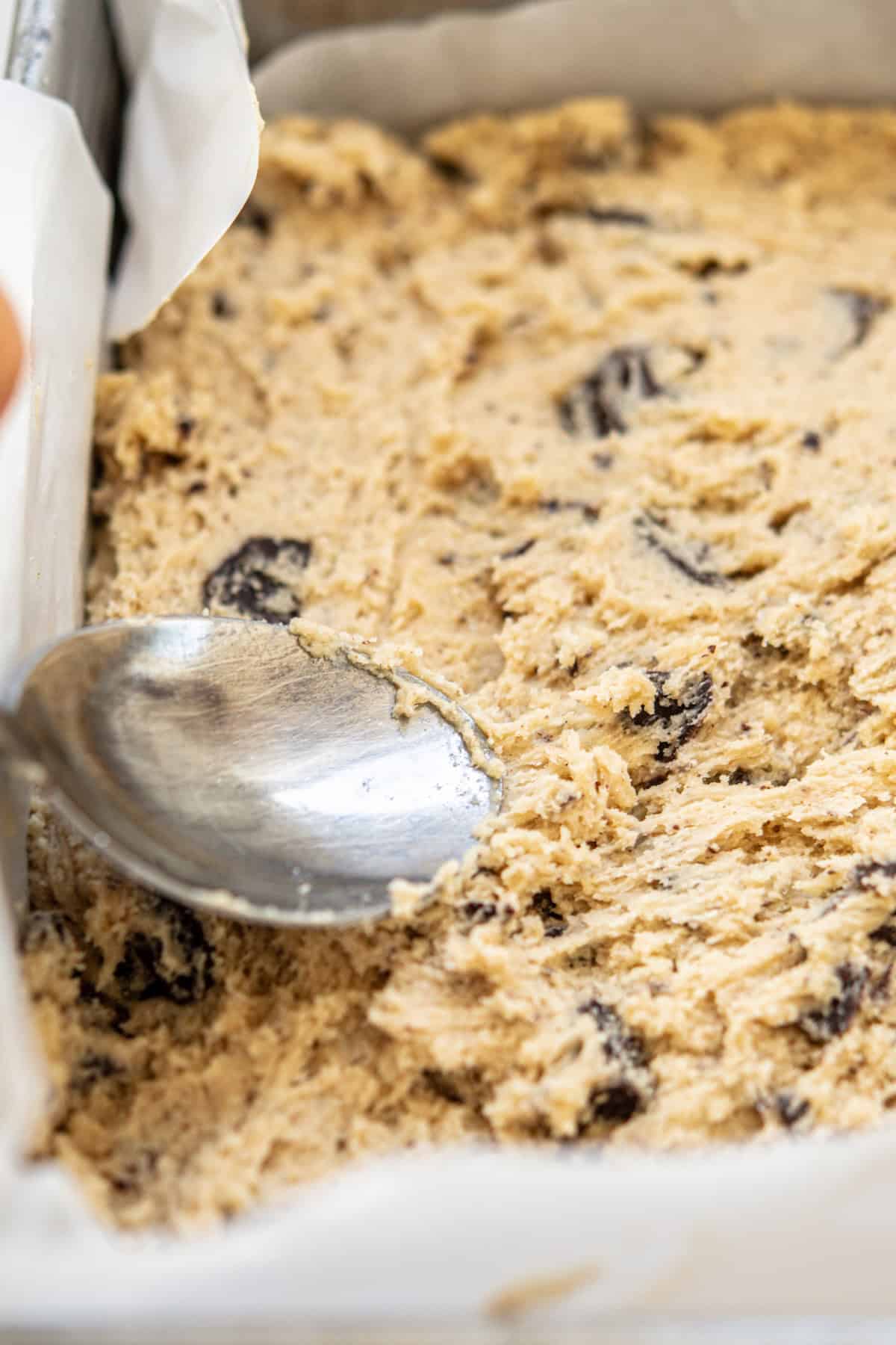 A close-up of sourdough cookie bars dough with chocolate chunks being spread evenly in a parchment-lined baking pan using the back of a spoon.