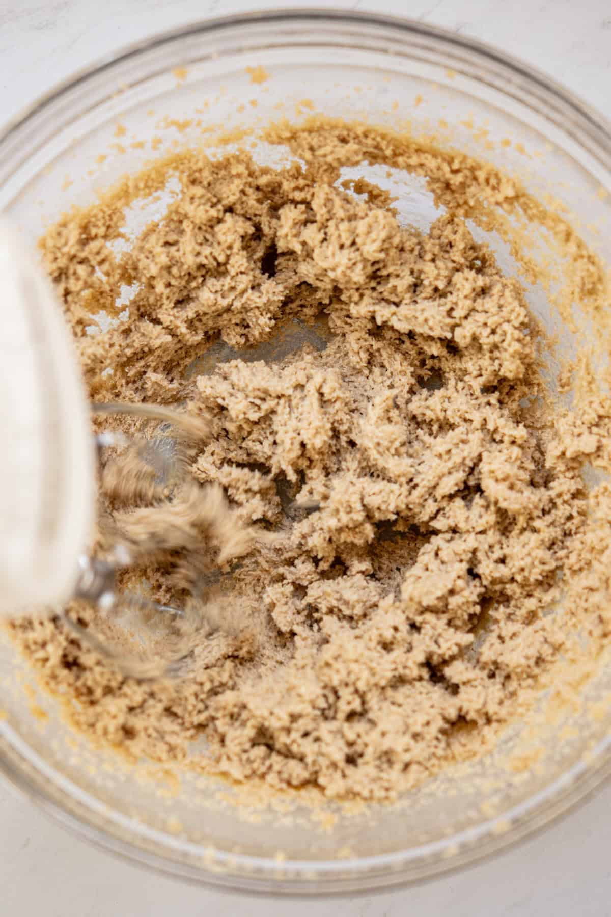 A glass mixing bowl containing creamed brown sugar and butter mixture for sourdough cookie bars is being mixed with a hand mixer on a light surface.