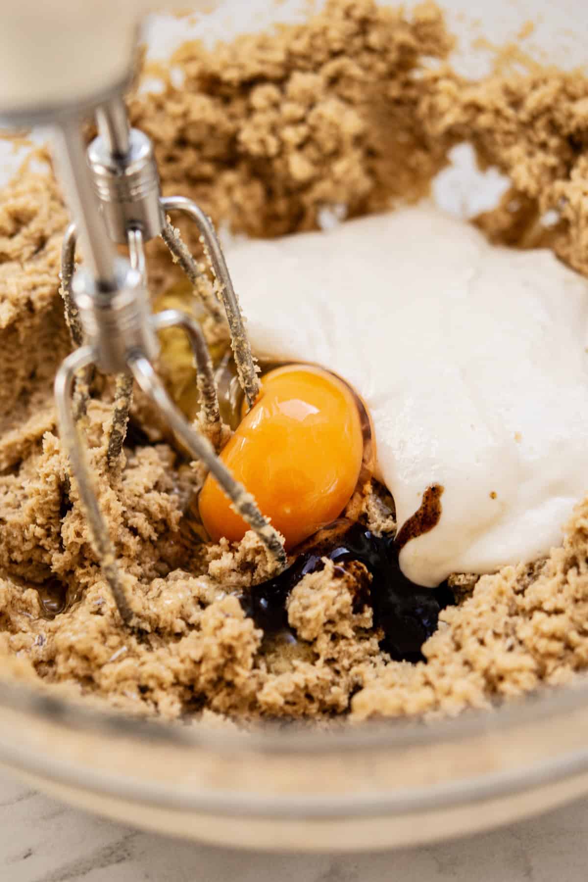 A close-up of an electric hand mixer beating sourdough cookie bars dough in a glass bowl, with visible brown sugar, an egg yolk, vanilla extract, and sour cream on top of the mixture.