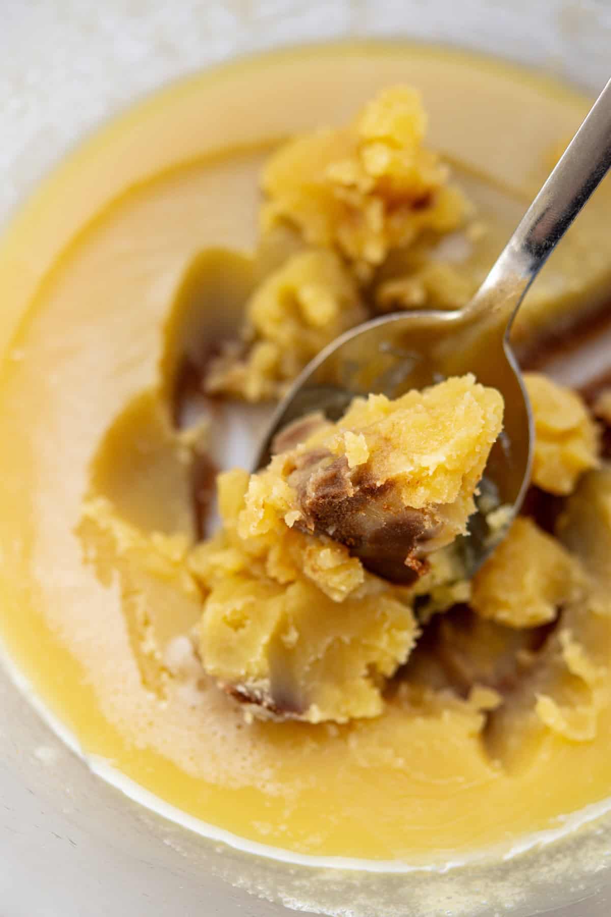A close-up of a metal spoon scooping thick, yellow-brown ghee from a glass container. The soft, creamy texture is perfect for baking treats like sourdough cookie bars, with some solidified and oily parts visible.