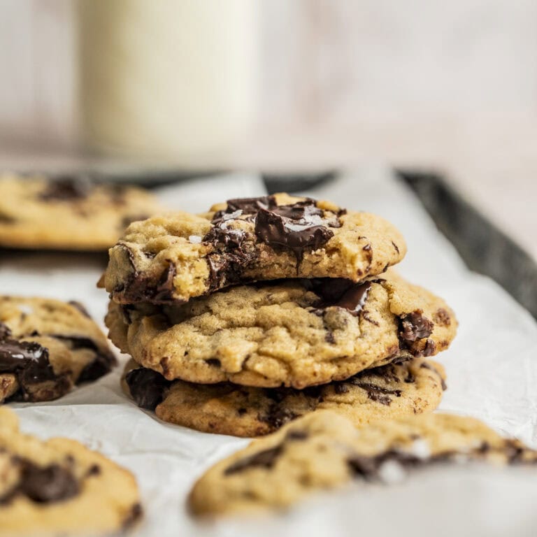 A stack of three Egg White Chocolate Chip Cookies with melted chocolate chunks sits on a baking sheet lined with parchment paper, with more cookies and a blurred glass of milk in the background.