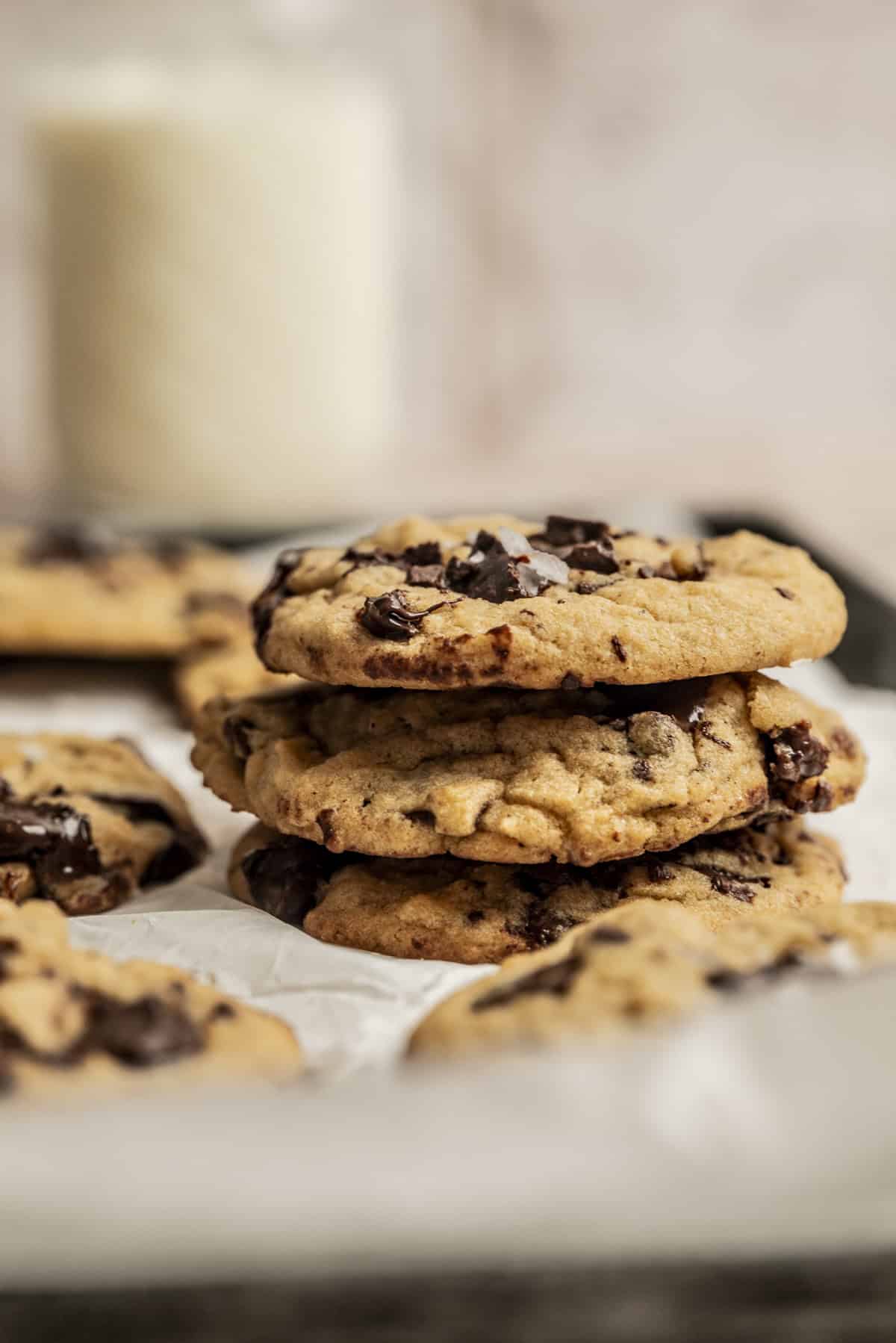 A stack of Egg White Chocolate Chip Cookies sits on parchment paper, with more cookies and a blurred glass of milk in the background.