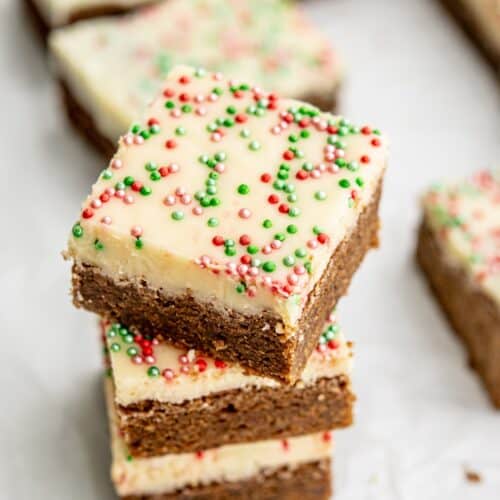 A stack of festive gingerbread bars topped with white icing and red, green, and white sprinkles on a white background. Several brownies are in the distance, creating a cheerful holiday presentation.