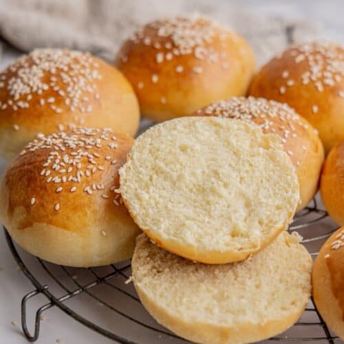 Freshly baked sesame seed sourdough buns are arranged on a cooling rack. One bun is cut in half, revealing a soft, fluffy interior. The golden-brown crust is speckled with sesame seeds.