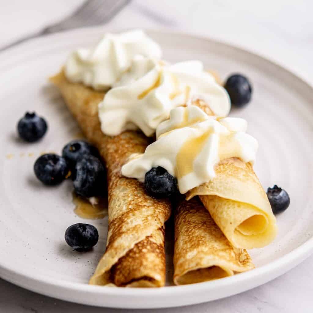Two rolled belgian pancakes topped with whipped cream and syrup, garnished with fresh blueberries, served on a white plate with a fork in the background.