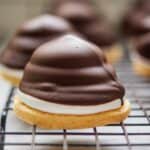 A close-up of a dessert featuring a crisp biscuit base topped with fluffy white marshmallow and covered in smooth dark chocolate. The dessert is resting on a wire rack, with similar treats blurred in the background.