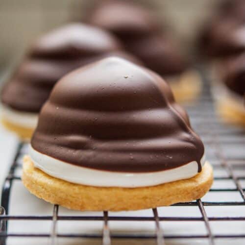 A close-up of a dessert featuring a crisp biscuit base topped with fluffy white marshmallow and covered in smooth dark chocolate. The dessert is resting on a wire rack, with similar treats blurred in the background.