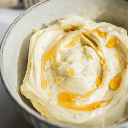 A bowl of creamy, whipped butter drizzled with golden honey. The textured swirls of butter and glistening honey create an appetizing appearance, reflecting light and adding a rich, inviting look to the dish.