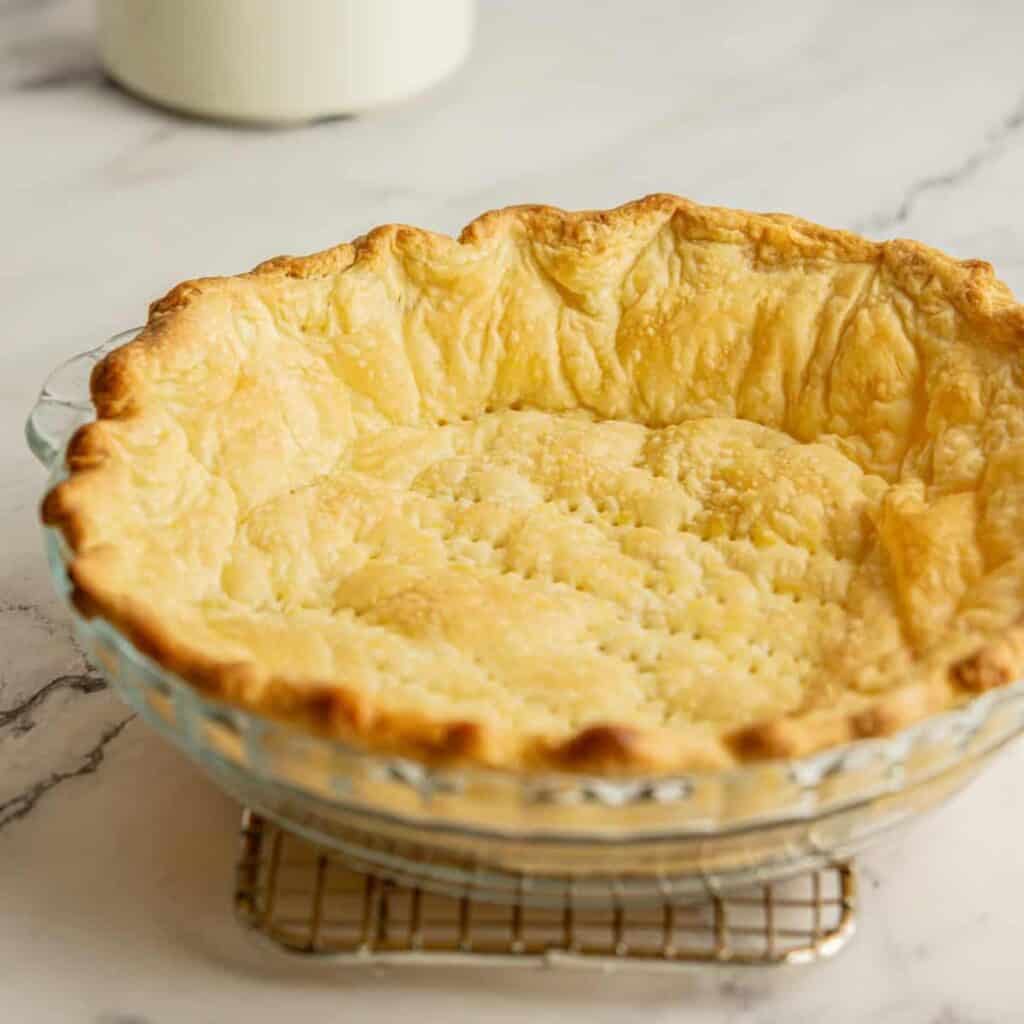 A golden brown pie crust in a glass dish sits on a cooling rack on a marble countertop. The crust is flaky and evenly baked, with a gently crimped edge.